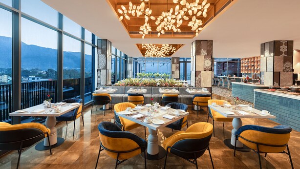 Un restaurante moderno con techos altos, amplios ventanales y vistas a la ciudad. Las mesas están dispuestas con manteles blancos y sillas amarillas.
