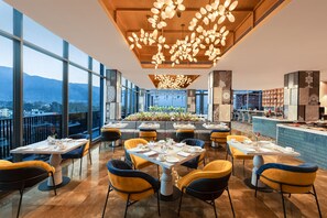 A modern restaurant with high ceilings, large windows, and a view of the cityscape. Tables are set with white tablecloths and yellow chairs. - Fortune Walkway Mall, Haldwani - Member ITC Hotels' Group (Haldwani)