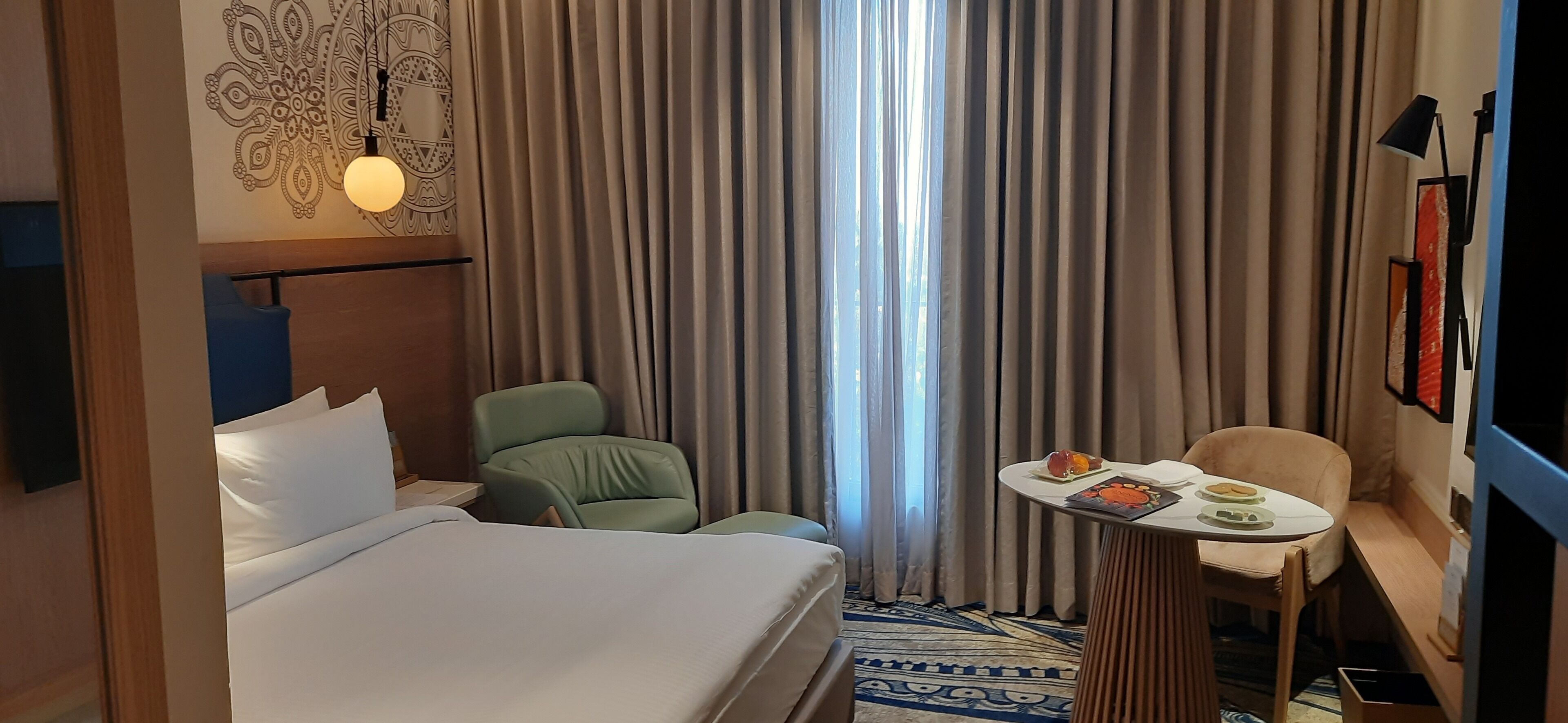 Une chambre d’hôtel avec un lit, un fauteuil vert, une petite table sur laquelle se trouve une assiette de fruits et un téléviseur fixé au mur.