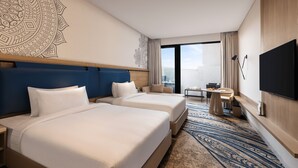 Habitación de hotel con dos camas, un mural grande en la pared, un televisor de pantalla plana, una mesita con una silla y vistas a la ciudad por la ventana.