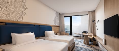 Habitación de hotel con dos camas, un mural grande en la pared, un televisor de pantalla plana, una mesita con una silla y vistas a la ciudad por la ventana.