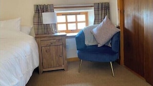 A bedroom with a bed, a nightstand with a lamp, a blue armchair, a window with curtains, and a wooden door. - The Stables Cottage (Darnick)