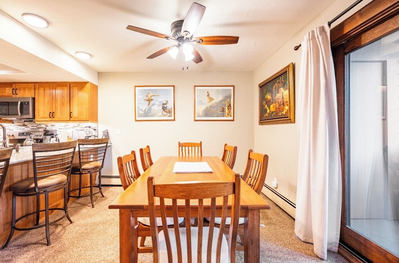 A dining area with a wooden table and chairs, a ceiling fan, and kitchen cabinets in the background.