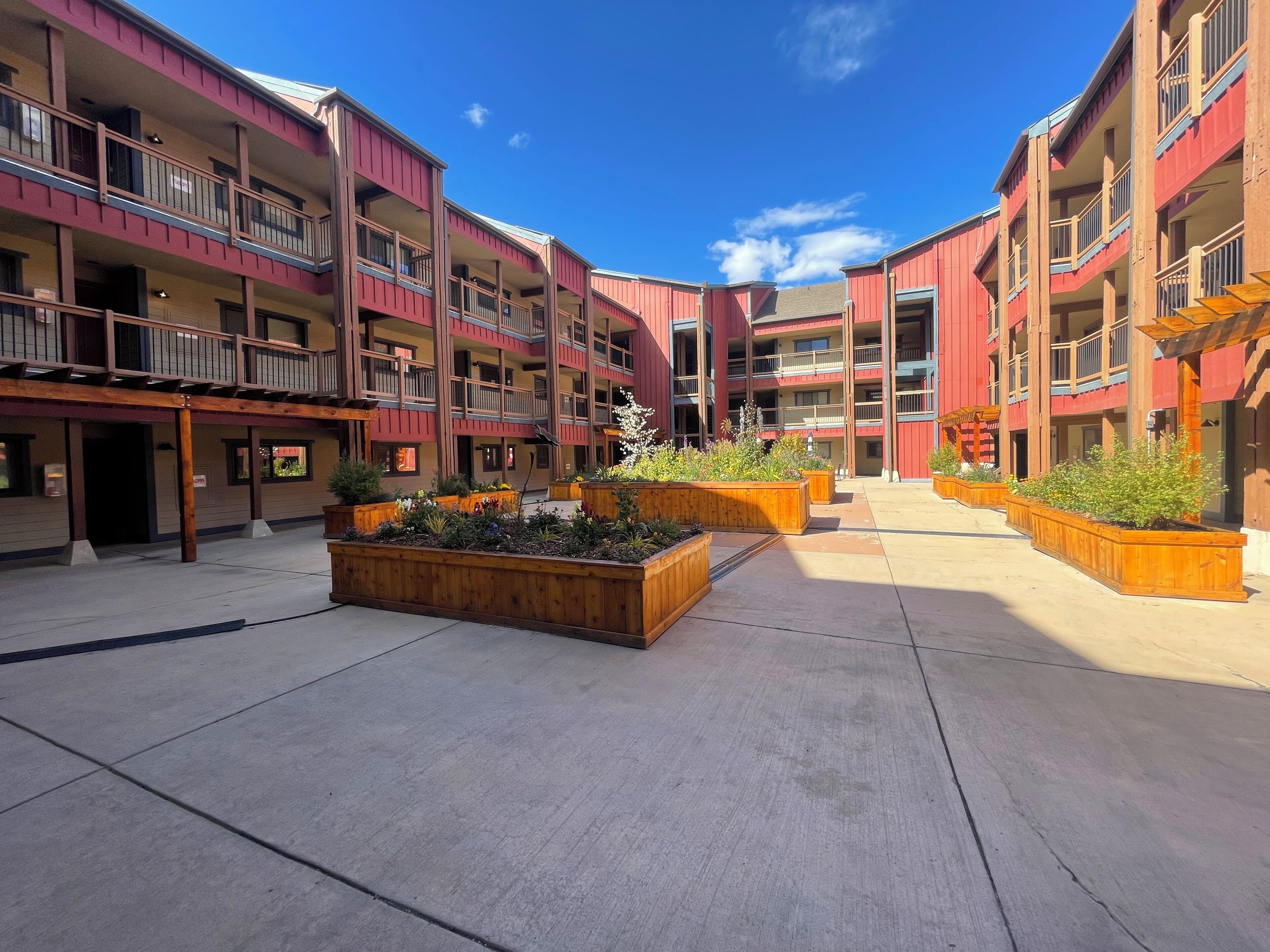 A courtyard with multi-story buildings, wooden planters with plants, and a clear blue sky.