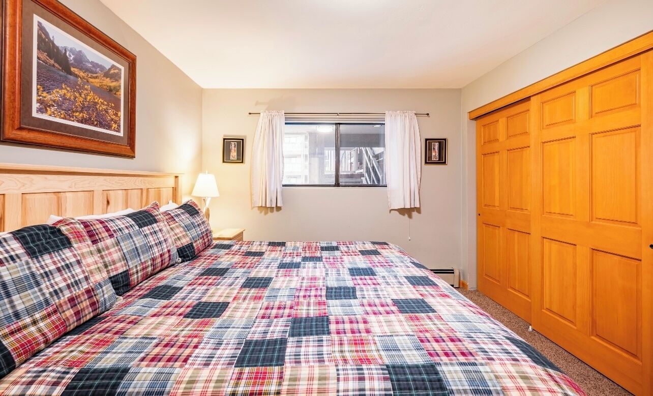 A bedroom with a plaid bedspread, a wooden headboard, a window with curtains, a framed picture on the wall, and a wooden wardrobe.