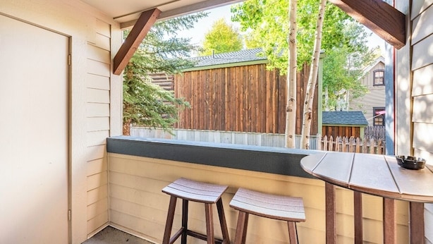 Un pequeño espacio al aire libre con una mesa de madera y dos taburetes, cerca de una valla de madera, con vista a una casa y árboles.