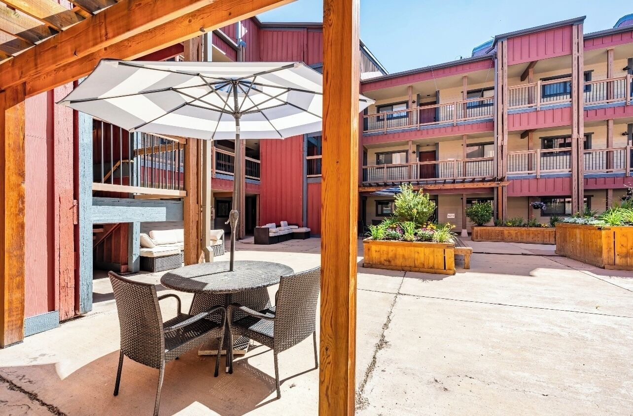 An outdoor patio area with a round table and chairs, a white umbrella, and wooden planters with greenery.