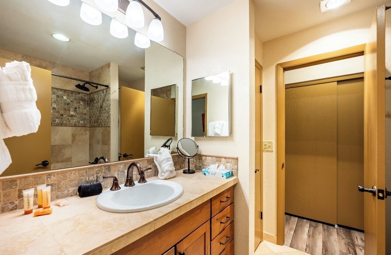 A bathroom with a large mirror, a sink, and a shower. There are towels and toiletries on the counter.