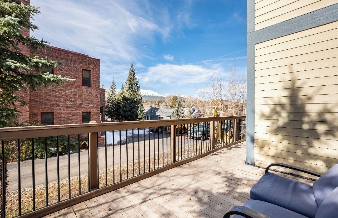 A wooden deck with a metal railing, a blue chair, and a view of a brick building and snow-capped mountains in the distance.