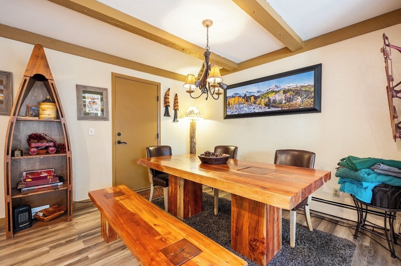 Un espace repas avec une table en bois, des bancs et des chaises. Un lustre est suspendu au-dessus de la table, et un tableau orne le mur.