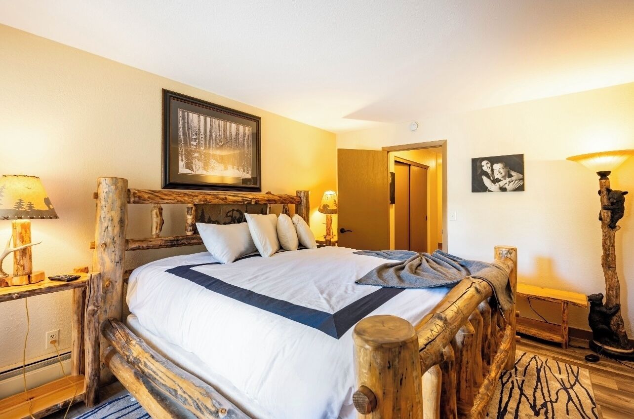 A wooden-framed bed with a white and blue bedspread, two bedside lamps, a framed picture above the bed, and a small bench with a lamp beside the bed.