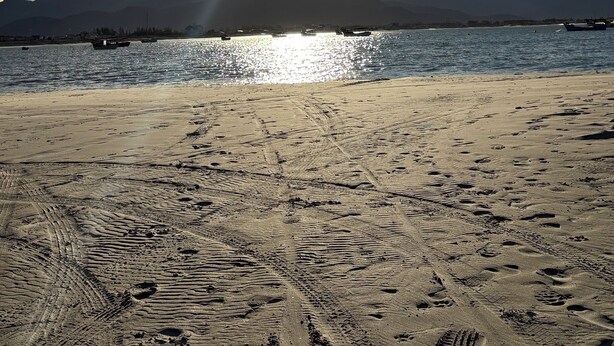 Ein Strand mit Fußabdrücken und Spuren von Reifen im Sand, ein Gewässer, Boote und Berge im Hintergrund.