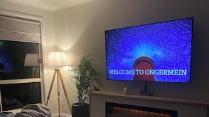 A modern living room with a flat-screen TV displaying "WELCOME TO ONERMEIN" on a wall-mounted screen, a fireplace below, a floor lamp, and a bean bag chair. - Ongermein, double story beach house a stone throw from the Indian Ocean.  (Port Macdonnell)