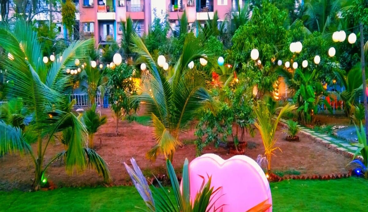 A lush garden with palm trees and string lights, a pink heart-shaped decoration, and a building with balconies in the background.
