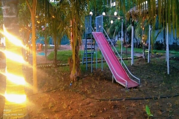 A playground with a red slide and a blue climbing structure, surrounded by palm trees and string lights. - Starlight Resort (Alibag)