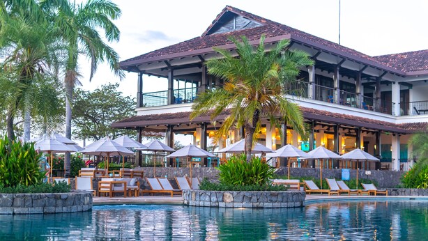Een resort met een zwembad, ligbedden, parasols en een gebouw met een pannendak.