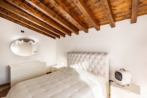 A bedroom with a tufted headboard, a round mirror, a bedside table with a lamp, and a portable heater.