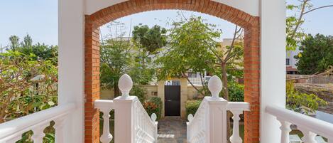 Un espacio al aire libre cubierto con un arco de ladrillo que conduce a una puerta, rodeado de barandillas blancas y follaje.