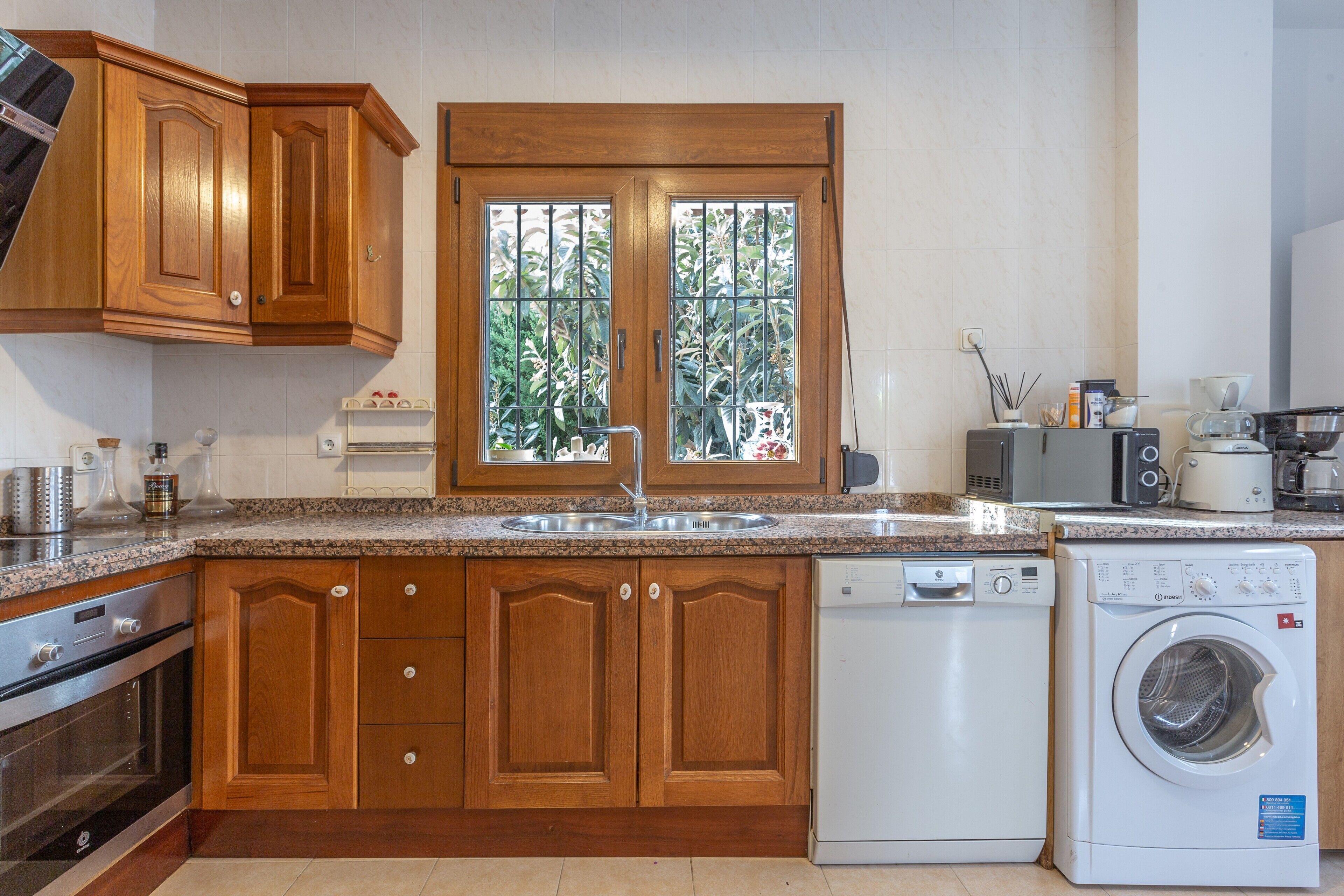 A kitchen with wooden cabinets, a built-in dishwasher, a washing machine, and a window with a view of greenery.