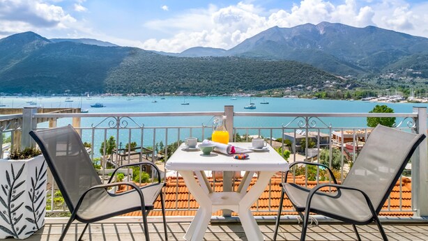 A balcony with two chairs, a table with a pitcher of orange juice, and a scenic view of a lake with boats and mountains. - Apartment 'Chakalaka - Suite with Sea View' with Sea View, Wi-Fi and Air Conditioning (Leukada)