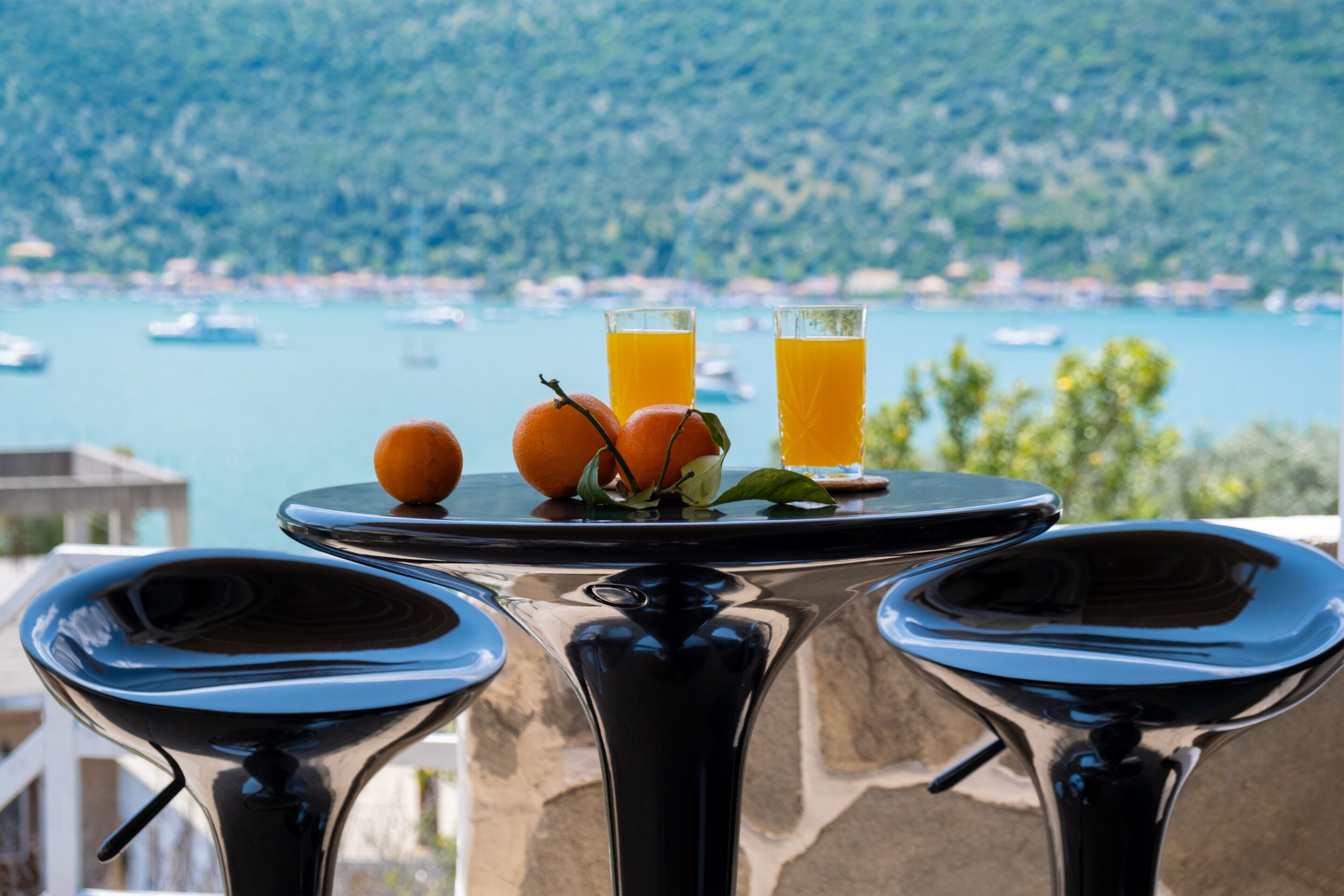 Una mesa con dos vasos de zumo de naranja y dos naranjas, con vistas a un cuerpo de agua y colinas.