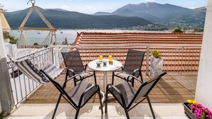 Een terras met uitzicht op bergen en water, met een hangmat, twee stoelen, een tafel met drankjes en een potplant.