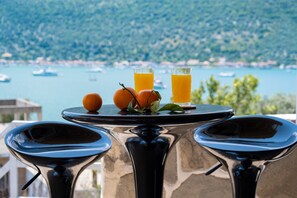 A table with two glasses of orange juice and two oranges, set against a scenic view of a body of water and hills.