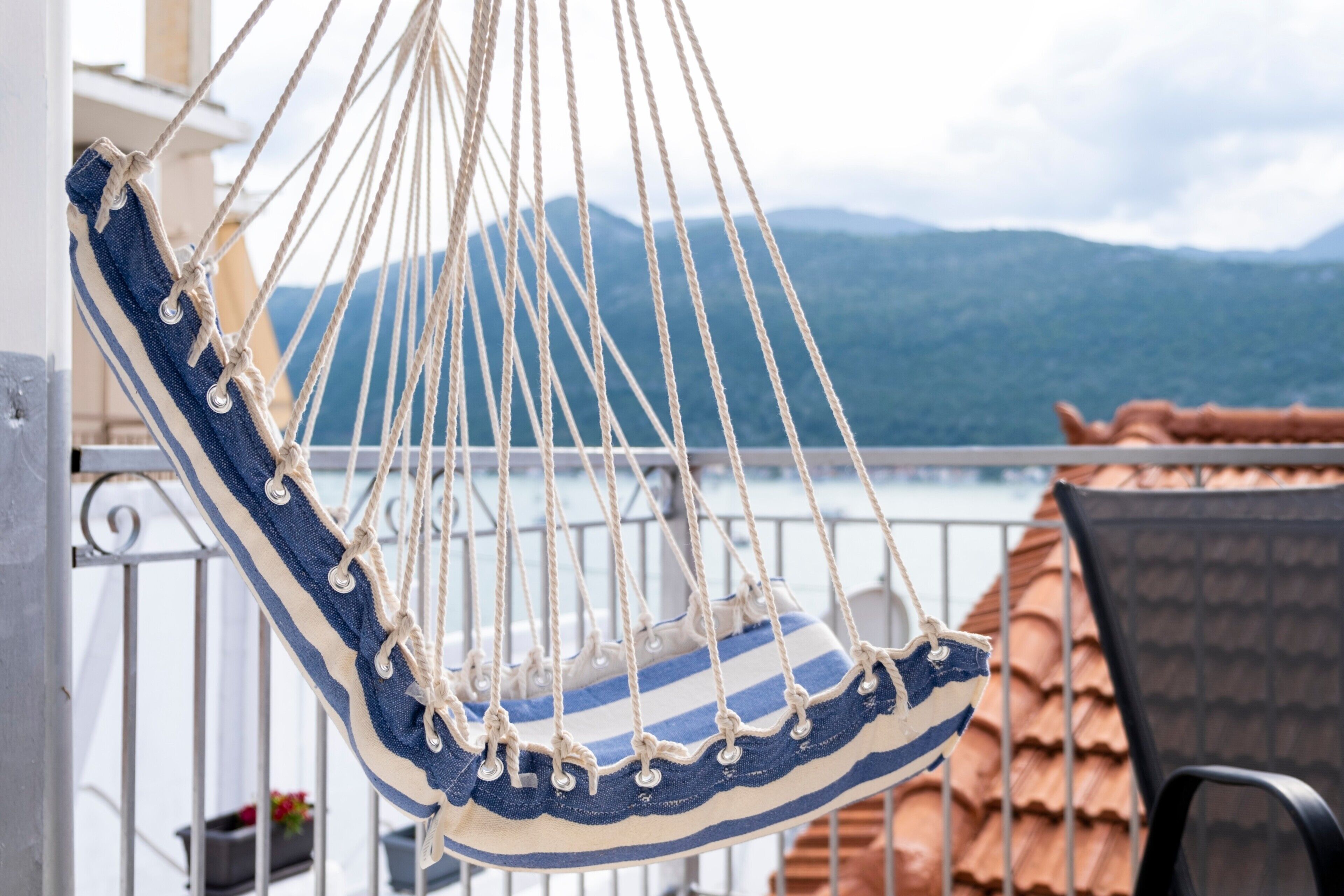 Un fauteuil suspendu en forme de hamac, avec un tissu à rayures, fixé par des cordes, sur un balcon doté d’une rambarde en métal et offrant une vue sur les montagnes.