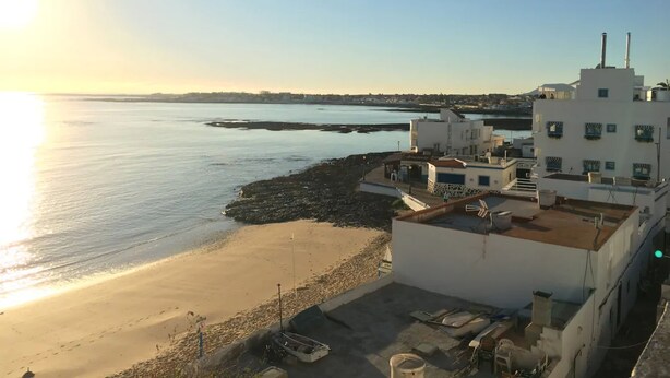 Un edificio frente al mar con terraza en la azotea, una playa arenosa y un mar tranquilo.