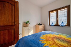 A bedroom with a wooden wardrobe, a bed with a sun-patterned quilt, a small wooden dresser, a window with curtains, and a plant on the dresser. - Holiday Home "Villa Pitti" with Sea View, Terraces & Wi-Fi (Castelsardo)