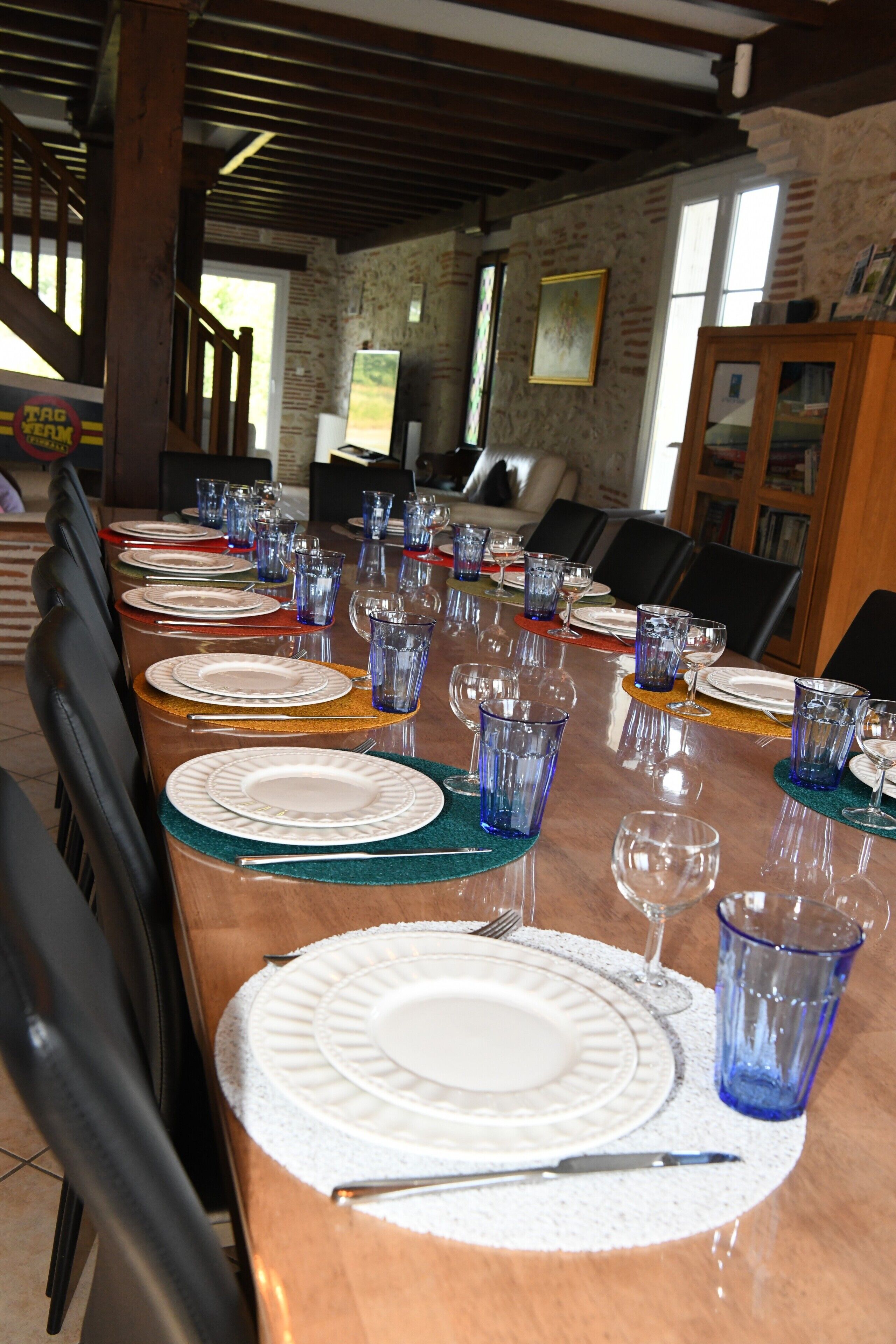 Une longue table à manger, dressée avec des assiettes, des verres et des couverts, entourée de chaises dans une pièce aux poutres apparentes et aux murs en briques.
