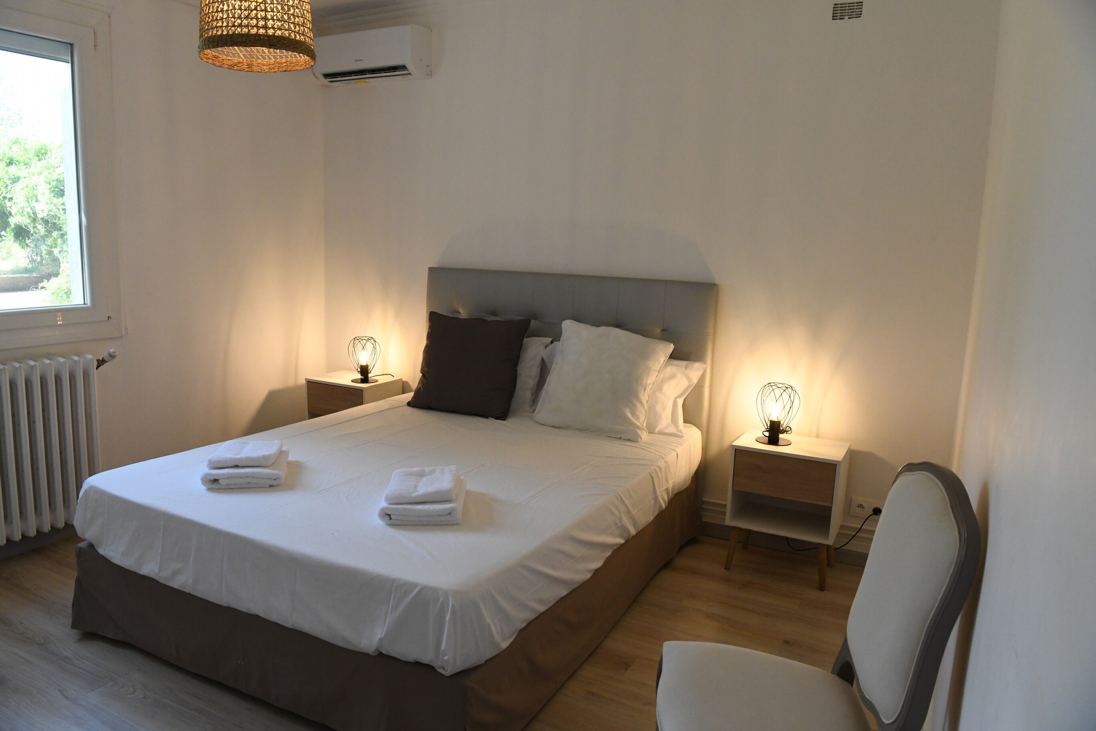 Uma cama bem arrumada com lençóis e toalhas brancos, uma mesa de cabeceira com abajur, uma cadeira e uma janela com vista para o verde.