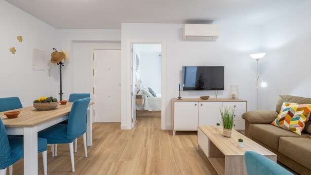 Un salon moderne comprenant un canapé, un téléviseur, un coin repas avec des chaises et une table, ainsi qu’un coin cuisine avec des placards et un plan de travail.