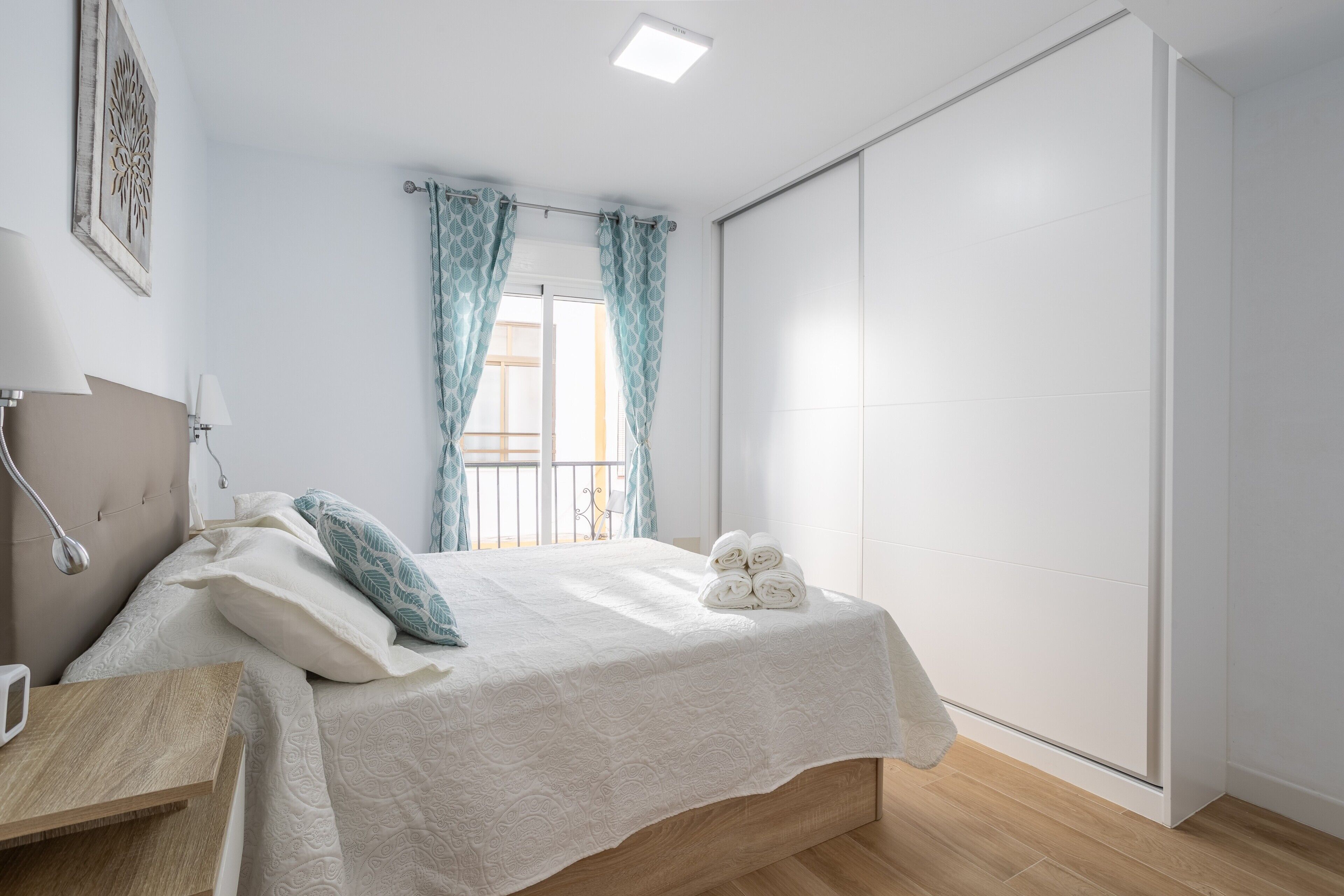 Una camera da letto con un letto, comodino, lampada, tende e vista sull'esterno.