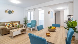 Un salon moderne comprenant un canapé, des fauteuils, une table basse et une table à manger avec des chaises. On aperçoit également un coin cuisine à travers une porte ouverte.