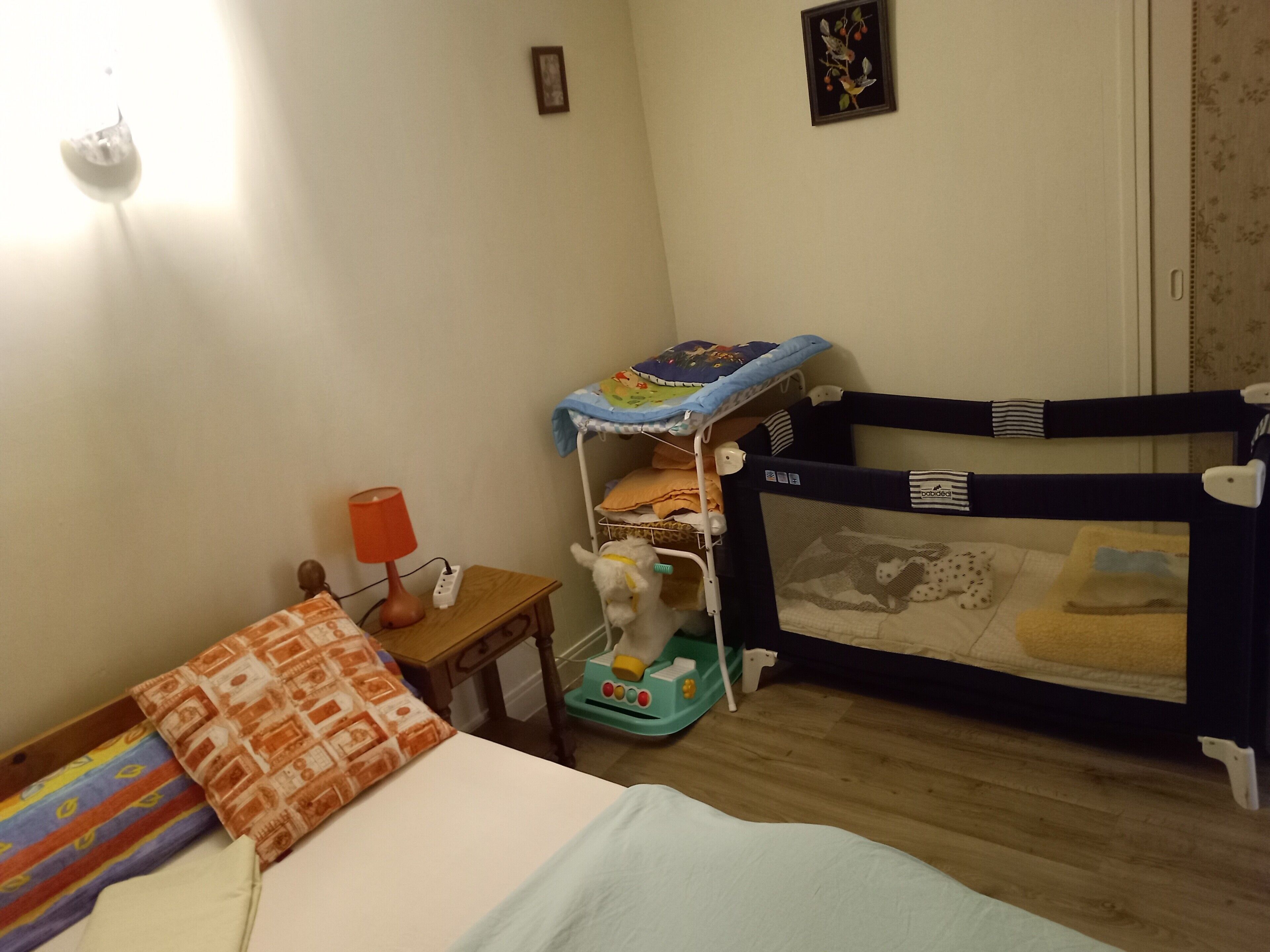 Quarto de criança com berço, uma pequena mesa com abajur, cama com manta estampada e um quadro na parede.