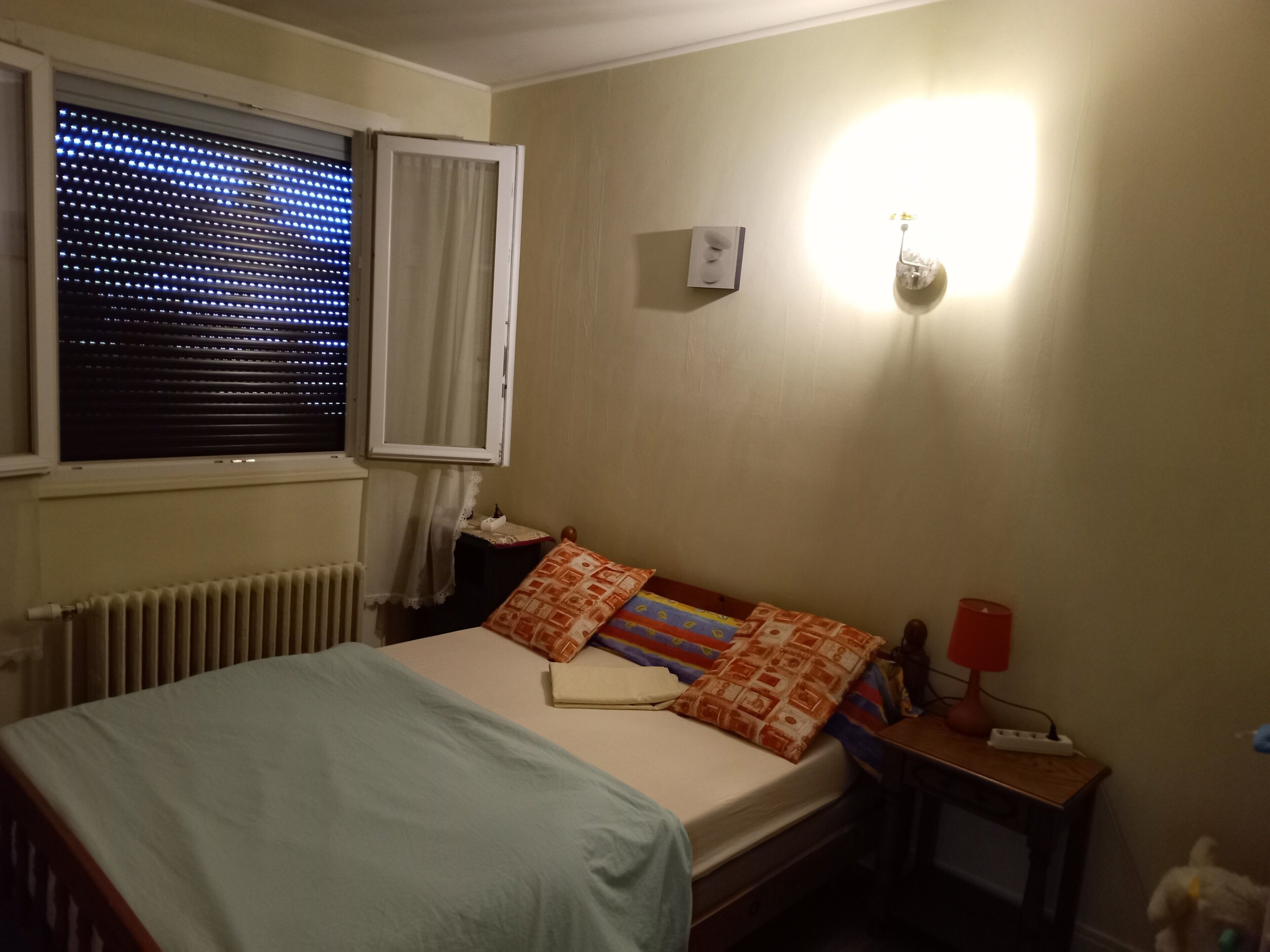 Une chambre à coucher comprenant un lit, une table de chevet, une lampe, une fenêtre avec des stores et un luminaire fixé au mur.
