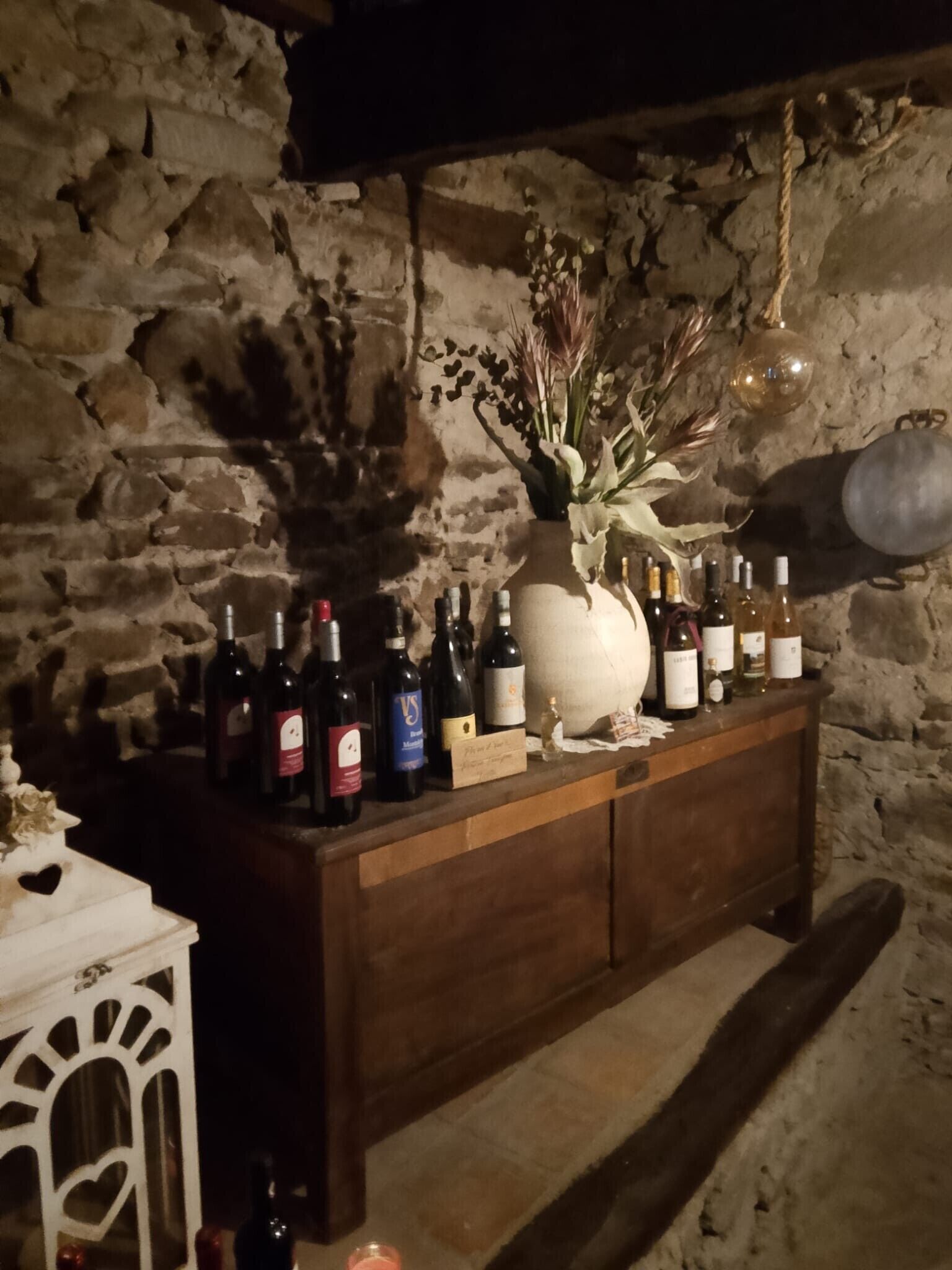 Un intérieur avec un mur en pierre et un bar en bois où sont exposées des bouteilles de vin, ainsi qu’un vase contenant des fleurs séchées.
