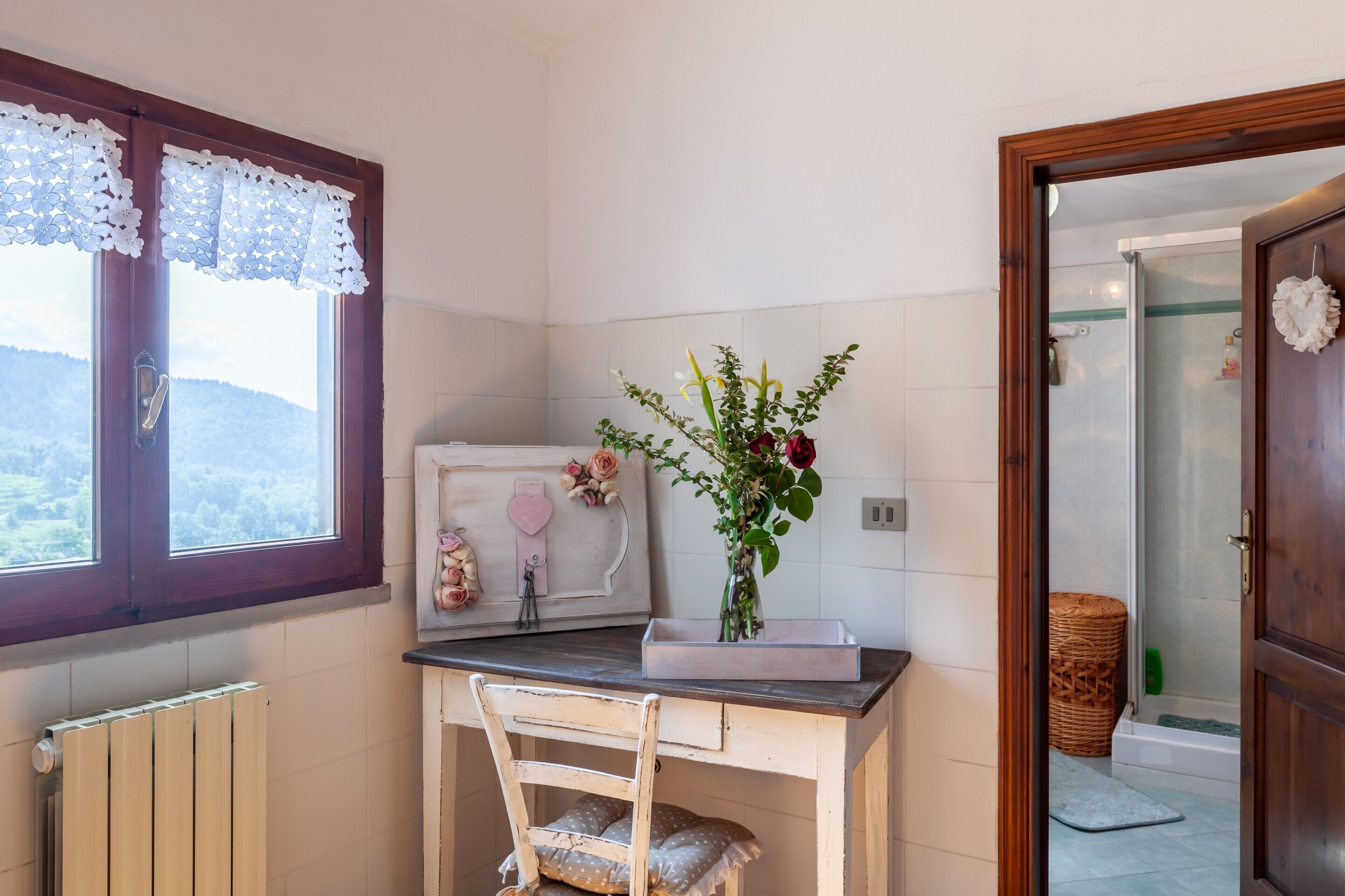 Een kleine keuken met een houten stoel, een vaas met bloemen, een raam met kantgordijnen en uitzicht op de omgeving.