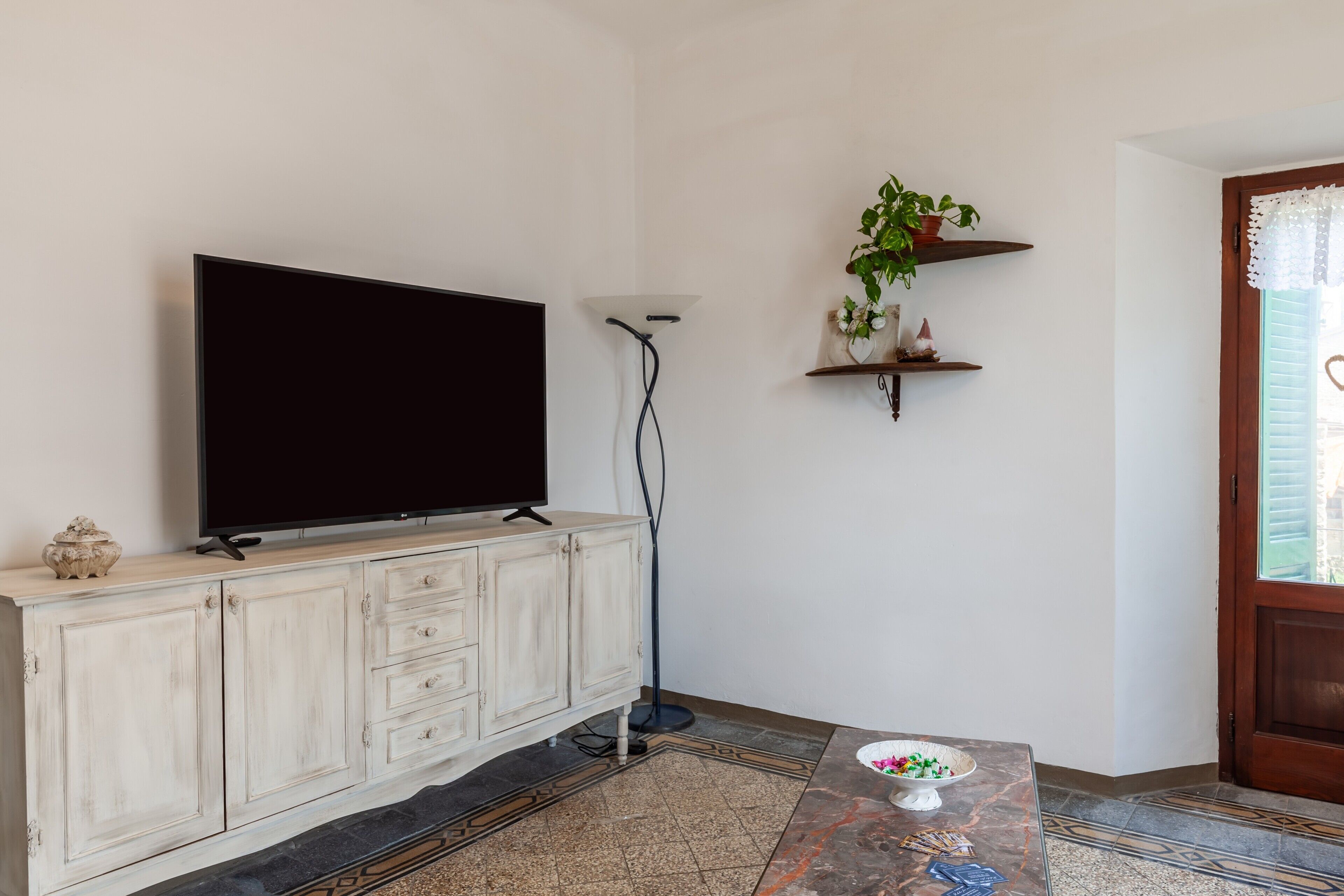 Una sala de estar con un televisor de pantalla plana sobre un mueble bajo, una lámpara de pie, una planta en maceta y una mesita con un cuenco.