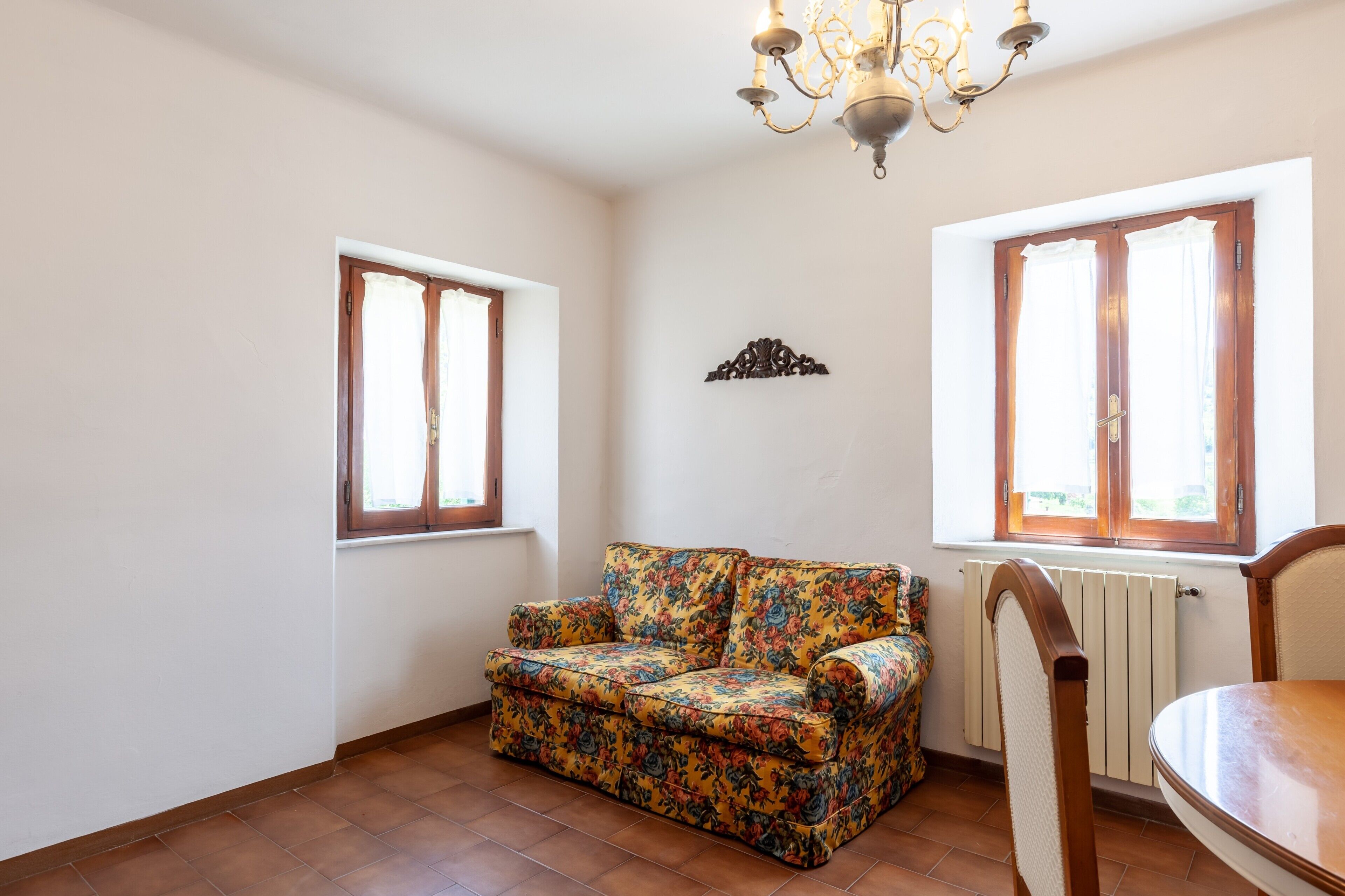 A room with a floral sofa, a dining table, a chandelier, and two windows with curtains.