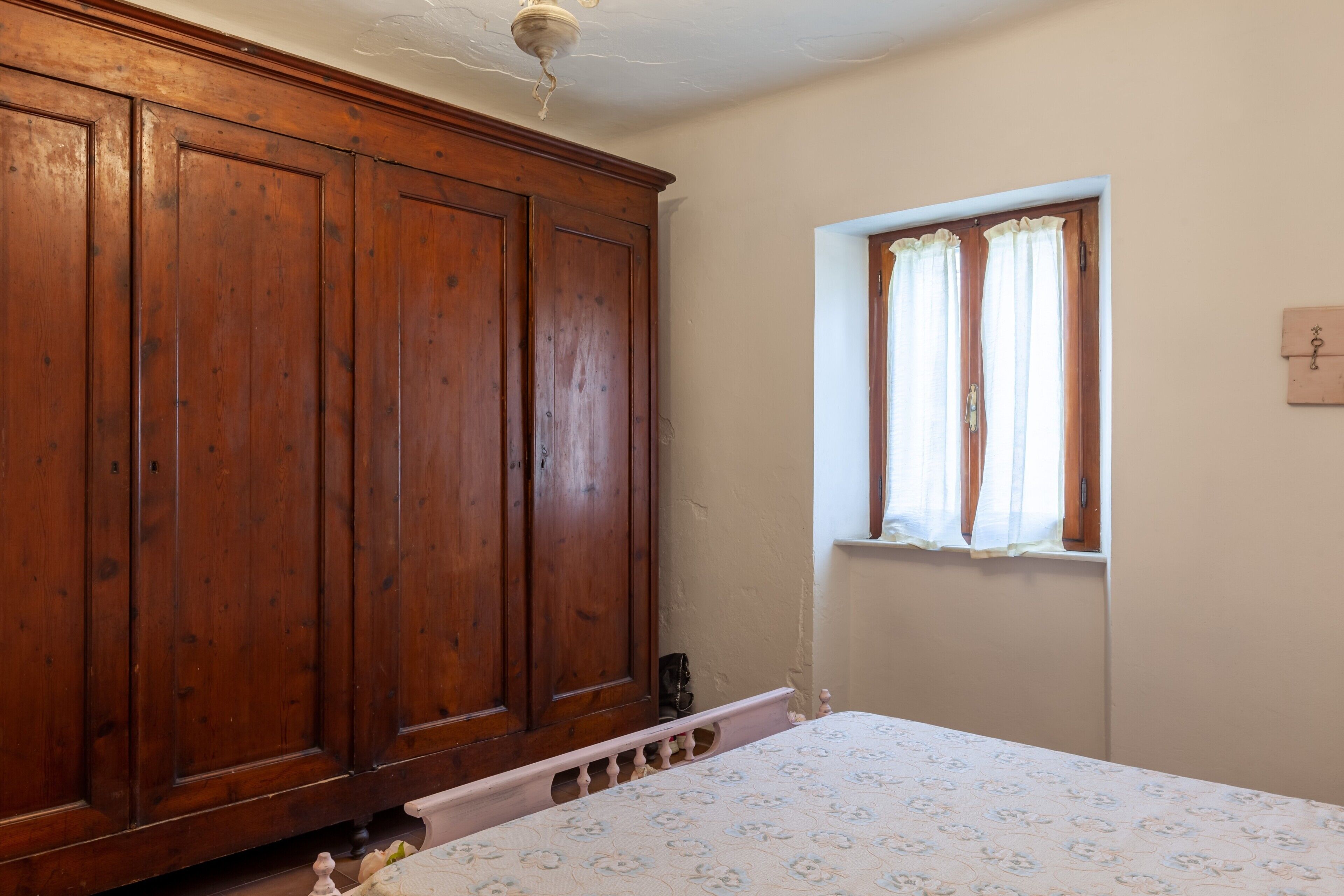 Ein Zimmer mit einem Holzschrank, einem Bett mit floralem Überwurf, einem Fenster mit Vorhängen und einem Lichtschalter an der Wand.