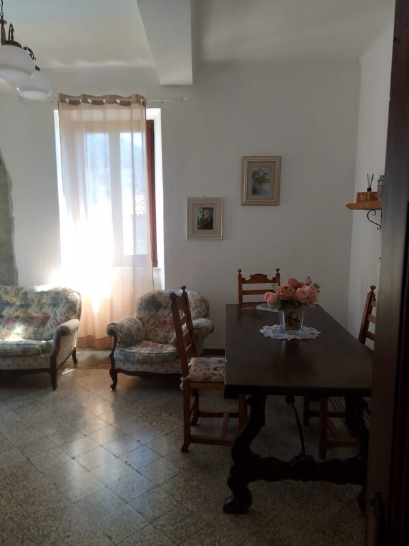 A dining area with a wooden table, two chairs, a floral sofa, and a window with curtains.