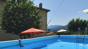 Piscina com água azul cristalina, rodeada por um deck branco, um guarda-sol vermelho e vista para montanhas ao fundo.