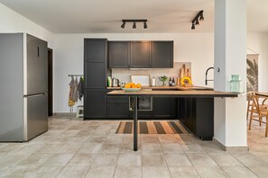 Une cuisine moderne avec des meubles de couleur sombre, un réfrigérateur en acier inoxydable, une table en bois et un sol carrelé.