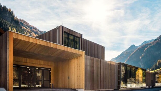 A modern building with a wooden facade and large windows, set against a mountainous backdrop.
