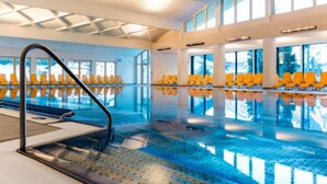 An indoor swimming pool with clear blue water, yellow lounge chairs, and large windows allowing natural light.