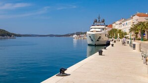 Ein Yachthafen mit einer großen Yacht an der Anlegestelle, einer gepflasterten Uferpromenade und Gebäuden mit roten Dächern.
