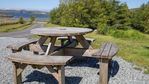 Une table de pique-nique en bois, avec des bancs, posée sur du gravier, entourée de verdure et offrant une vue sur un lac et des collines.