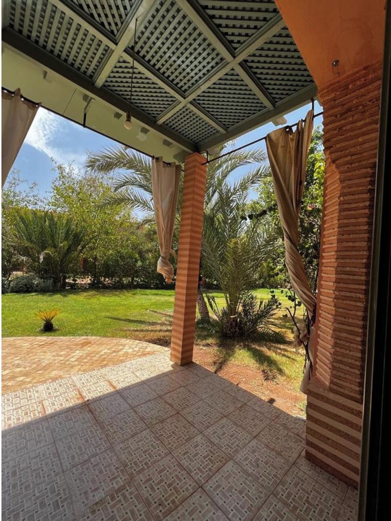 Un patio techado con piso de baldosas, palmeras y vista a un césped bien cuidado.